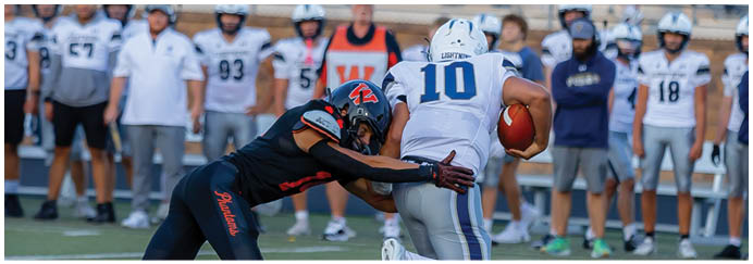 A person in a dark sports uniform is tackling another person in a white sports uniform who is holding a football on a field with other people standing in the background. Description generated by AI