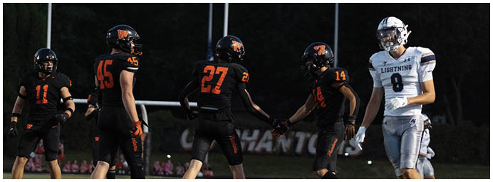 Five people wearing football uniforms stand on a field at dusk, with four in black and orange uniforms and one in a white and blue uniform. Description generated by AI
