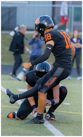 Two people in black football uniforms with orange accents are on a field, with one holding the ball in place while the other prepares to kick it. Description generated by AI