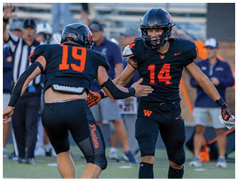 Two people in black and orange football uniforms are on a field with one reaching out to the other while a referee and other players stand in the background. Description generated by AI