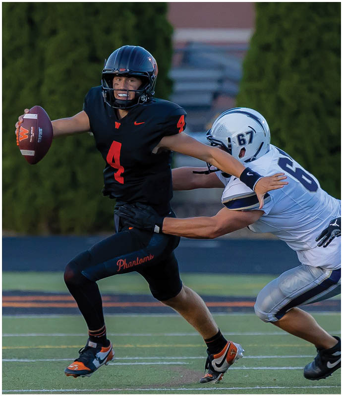 A person wearing a black football uniform with the number 4 is running on a field while holding a football and being tackled by another player in a white uniform. Description generated by AI