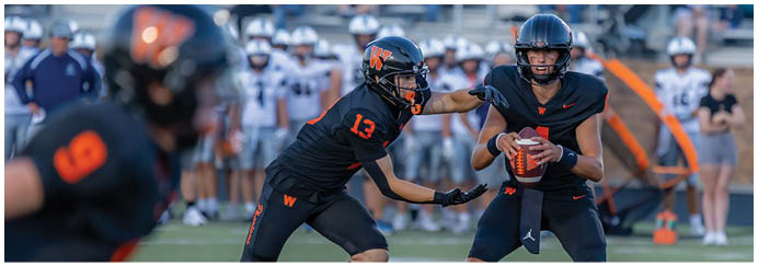 Two people in black sports uniforms are playing football on a field, with one holding the ball and the other reaching out to block or tackle. Description generated by AI