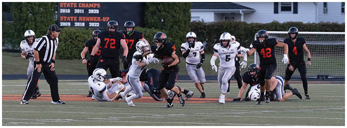 A group of people in football uniforms are actively playing on a field with a scoreboard in the background displaying information about West De Pere Football. Description generated by AI