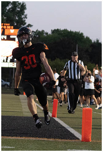 A person in a white football uniform is preparing to throw a pass while two opposing players in red and black uniforms attempt to block the throw on a football field. Description generated by AI