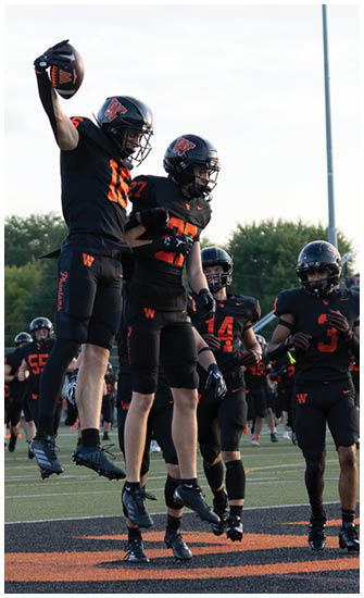 Two people in black and orange football uniforms are jumping and celebrating on a field while others stand nearby. Description generated by AI