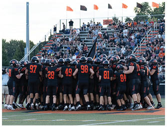 A group of football players in black and orange uniforms huddle together on a field with a crowd of spectators seated in bleachers behind them. Description generated by AI