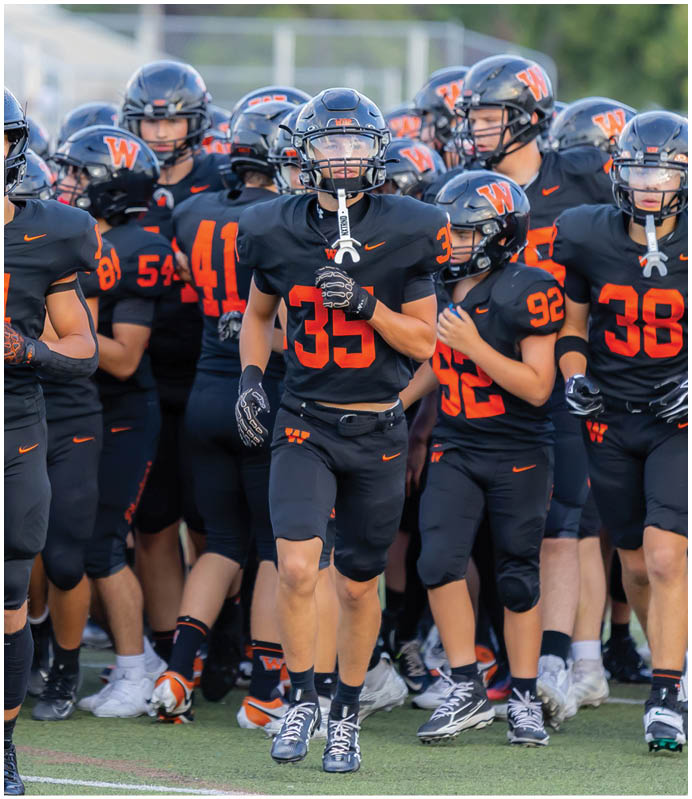 A group of people wearing black and orange football uniforms are running together on a sports field. Description generated by AI