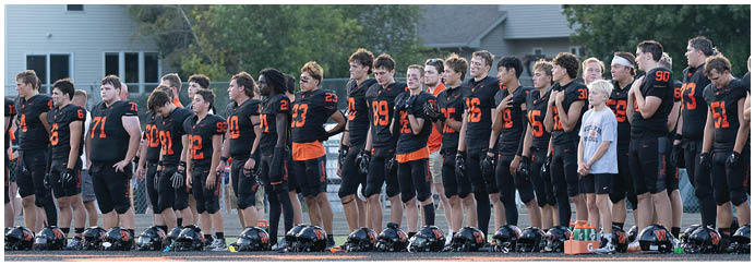 A group of people wearing black and orange football uniforms stand in a line outdoors, some with their hands over their hearts. Description generated by AI