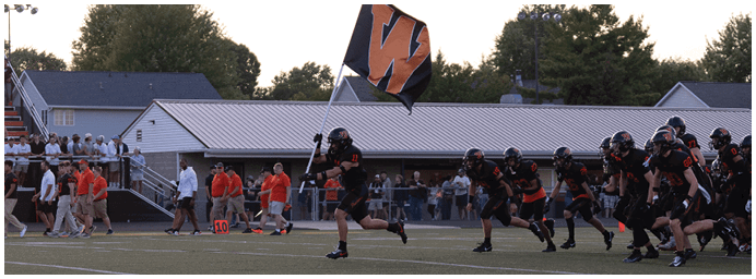 A group of people wearing black and orange sports uniforms are huddled together on a football field with a crowd seated in bleachers in the background. Description generated by AI