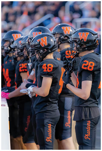 A person wearing a black football uniform with orange lettering and the number 28 stands on a track near a crowd in the background. Description generated by AI