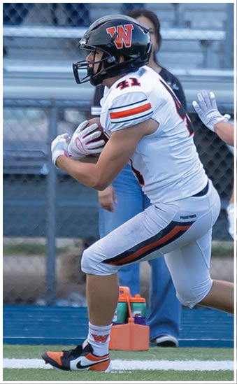 A person wearing a football uniform and helmet is holding a football and running on a field near a chain-link fence and bleachers. Description generated by AI
