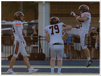 Two people in white football uniforms with black helmets stand on a field while another person in a similar uniform raises a leg in the background. Description generated by AI