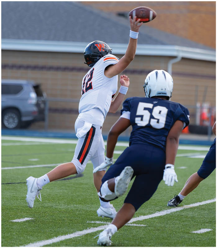 A person in a white football uniform is throwing a football while a person in a dark blue uniform with the number 59 runs toward them on a football field. Description generated by AI