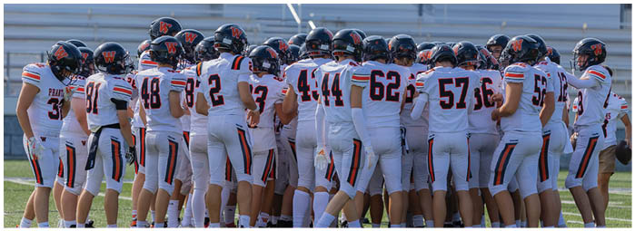 A group of people wearing white sports uniforms with black helmets and orange and black details are gathered closely together on a field. Description generated by AI