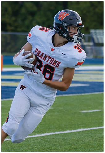 A person wearing a white football uniform with “Phantoms" on the jersey and a black helmet is running on a football field while holding a football. Description generated by AI