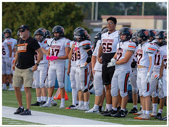 A group of people wearing football uniforms and helmets stand on the sidelines of a football field with one person in a black shirt and shorts gesturing. Description generated by AI