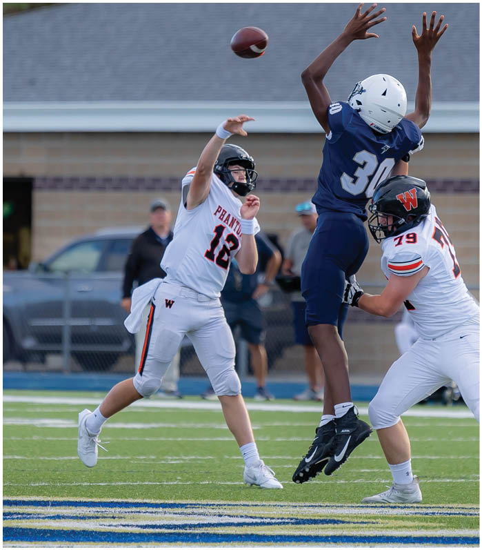 A person in a white football uniform is throwing a football while another person in a blue uniform jumps with arms raised and a third person in white attempts to block. Description generated by AI