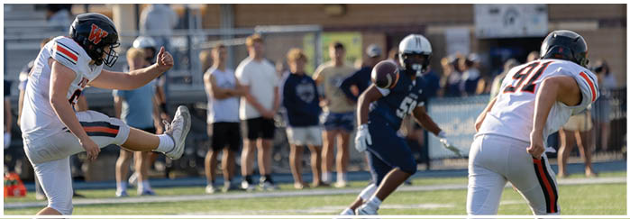 People in sports uniforms are playing football on a field with one person kicking the ball and others running. Description generated by AI