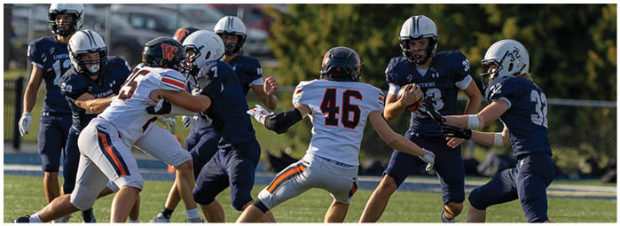 People wearing football helmets and uniforms are engaged in a play on a grassy field with trees and parked cars in the background. Description generated by AI