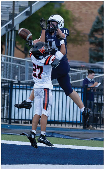 Two people in football uniforms are jumping and reaching for a football in midair during a game. Description generated by AI