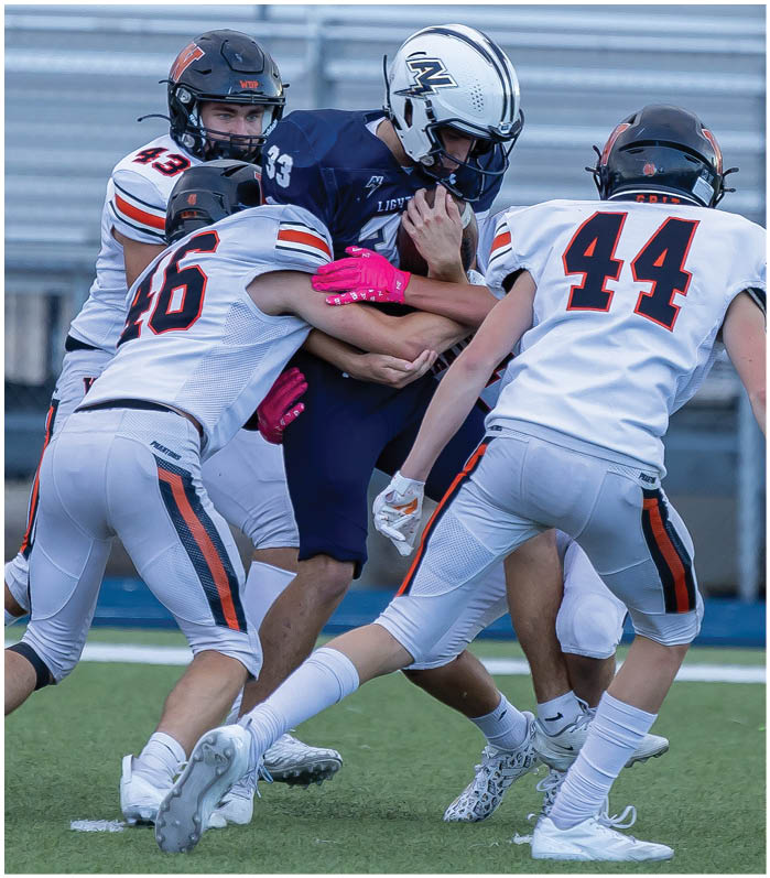 Three people in football uniforms are tackling a player in a dark blue uniform who is holding a football on a field. Description generated by AI