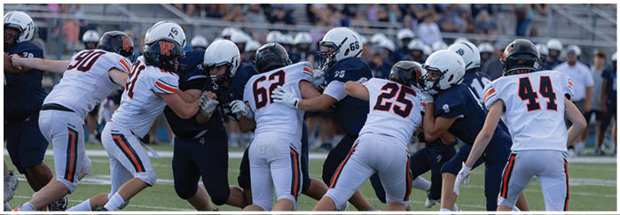 A group of people wearing football helmets and uniforms are engaged in a play on a football field with spectators visible in the background. Description generated by AI