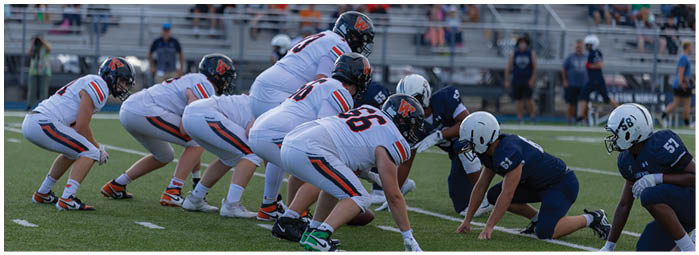Two opposing American football teams are lined up at the line of scrimmage on a green field with spectators in the background. Description generated by AI