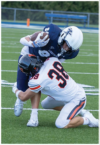 Two people in football uniforms are engaged in a tackle on a green field with one person holding the ball and the other attempting to stop them. Description generated by AI