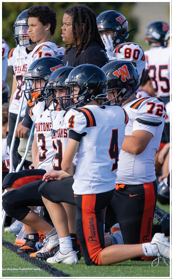 Several people in football uniforms with helmets are kneeling on a field while others stand behind them. Description generated by AI
