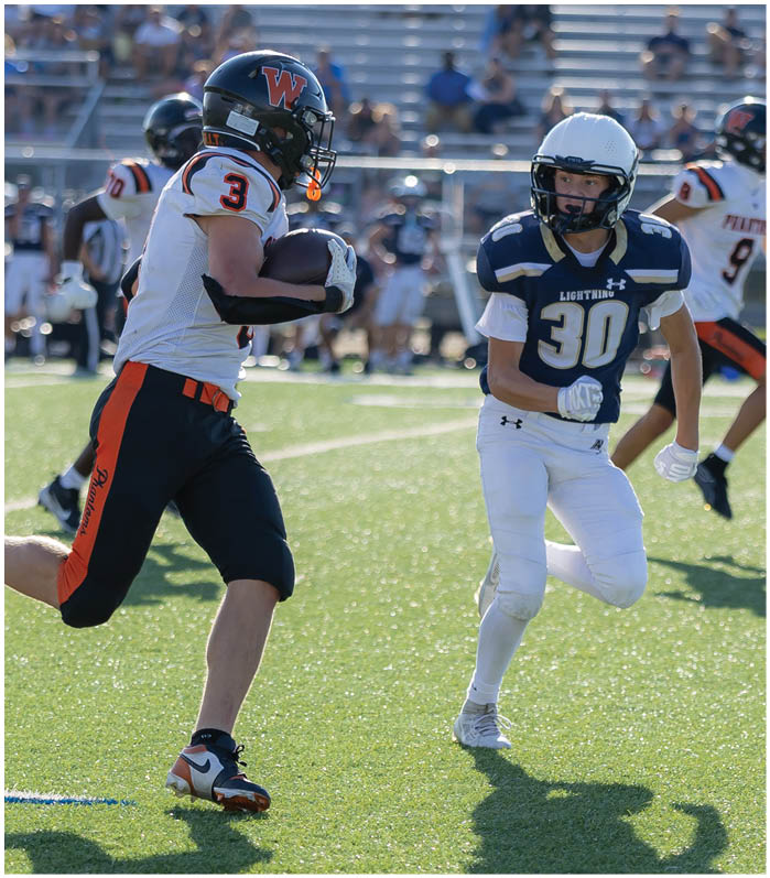 A person wearing black and orange athletic pants and cleats is running on a grassy sports field. Description generated by AI