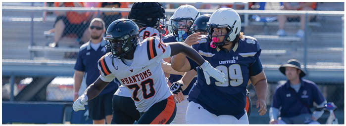 Two people in football uniforms are actively running on a field with one person wearing a white and orange jersey and the other wearing a blue and white jersey, while others and bleachers are visible in the background. Description generated by AI