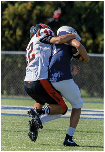 An adult wearing a blue jersey and white pants is holding a football while running on a grassy field with a chain-link fence and trees in the background. Description generated by AI