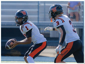 Two people wearing football helmets and uniforms are on a field with one holding a football and the other preparing to run. Description generated by AI