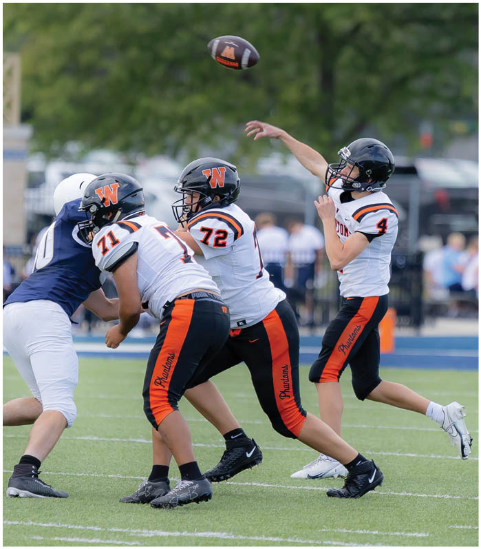 Three people in football uniforms and helmets are on a field with one person throwing a football while the others prepare to block. Description generated by AI