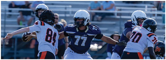 People wearing football helmets and uniforms are actively engaged in a game on a field with bleachers and spectators in the background. Description generated by AI
