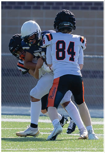 Three people wearing football uniforms and helmets are engaged in a play on a grassy field with a brick wall in the background. Description generated by AI