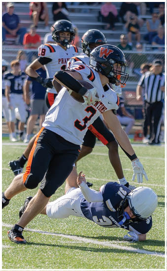 A person in a white and navy football uniform is falling to the ground while several people in white and black football uniforms with orange accents are reaching toward them on a sunny field. Description generated by AI