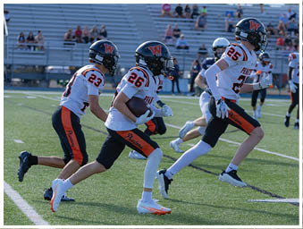 People in football uniforms are running on a field during a game, with one person holding the ball and others in motion behind. Description generated by AI