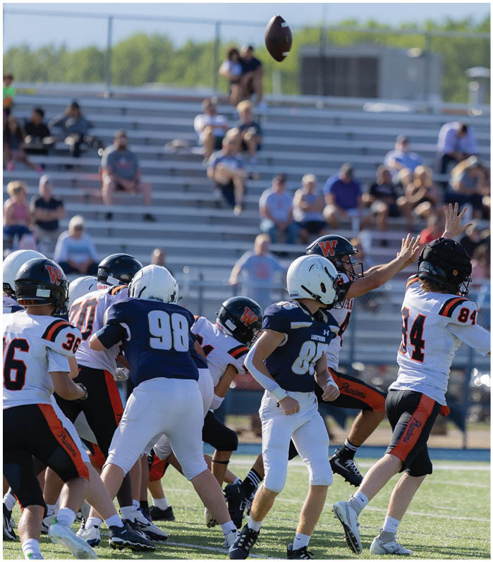 Several people in football uniforms are actively reaching for a football in midair on a field with bleachers and spectators in the background. Description generated by AI