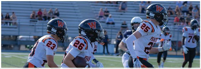 Three people in football uniforms with helmets are running on a field during a game with spectators in the background. Description generated by AI