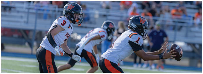 Three people in football uniforms are on a field with one person preparing to catch or receive a football. Description generated by AI