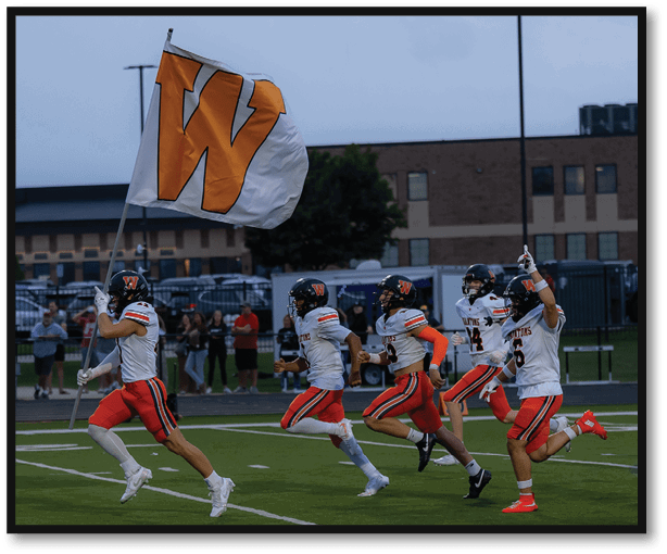 A group of people in football uniforms are running on a field with one person holding a large flag featuring a bold orange “W" against a white background. Description generated by AI