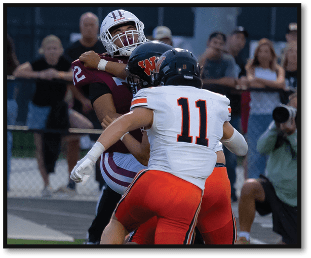 Several people in sports uniforms are actively engaged in a football game on a field with spectators in the background. Description generated by AI