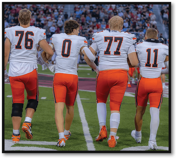Three people in football uniforms with numbers 76, 0, and 77 walk arm in arm on a football field toward a crowd in the stands. Description generated by AI
