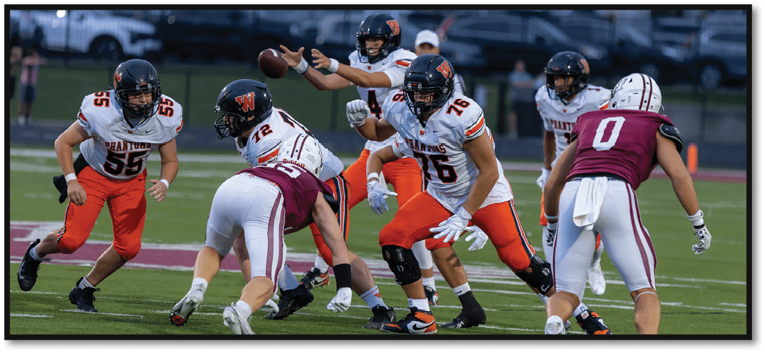 Players in football uniforms are actively engaged in a play on a field, with one player preparing to throw the ball while others block or move toward opponents. Description generated by AI