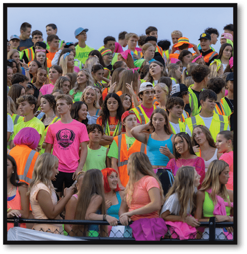A large group of people wearing colorful clothing and safety vests are gathered closely together, some standing and some leaning on a fence, engaging in conversation and looking in various directions. Description generated by AI