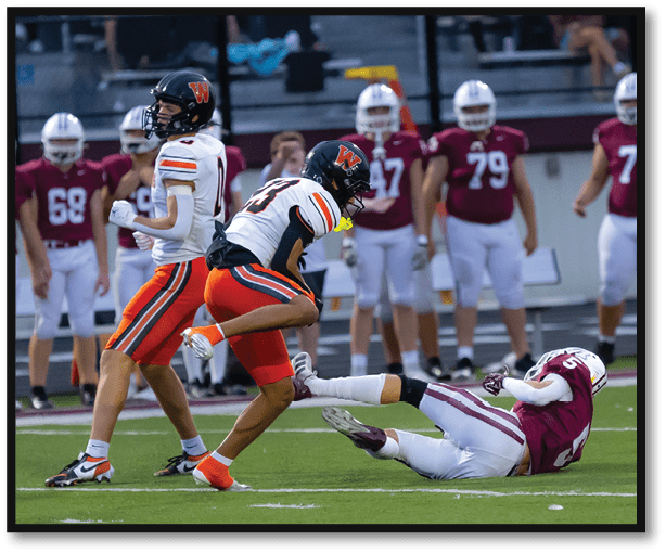 Two people in football uniforms are engaged in a play on a field with one person on the ground and the other mid-stride, while others in similar uniforms stand in the background. Description generated by AI