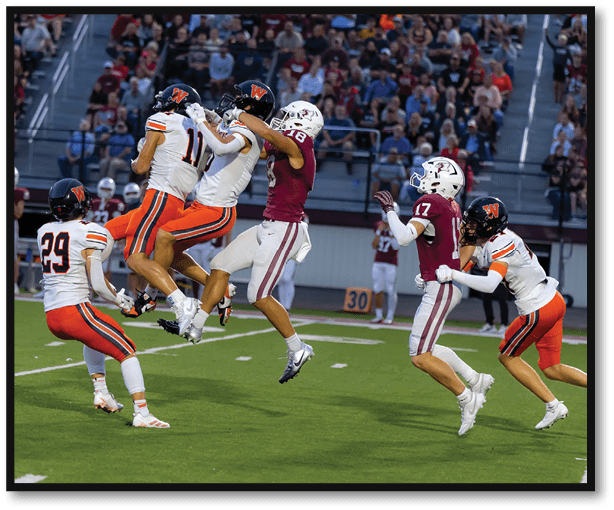Two teams of people in football uniforms are competing on a field with one player in white and orange jumping to catch the ball while a player in maroon attempts to block. Description generated by AI