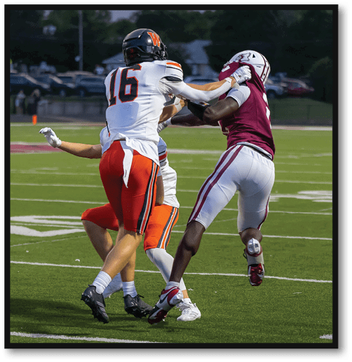 Four people in football uniforms are engaged in a play on a green field with one player in white and orange attempting to block another player in maroon and white. Description generated by AI