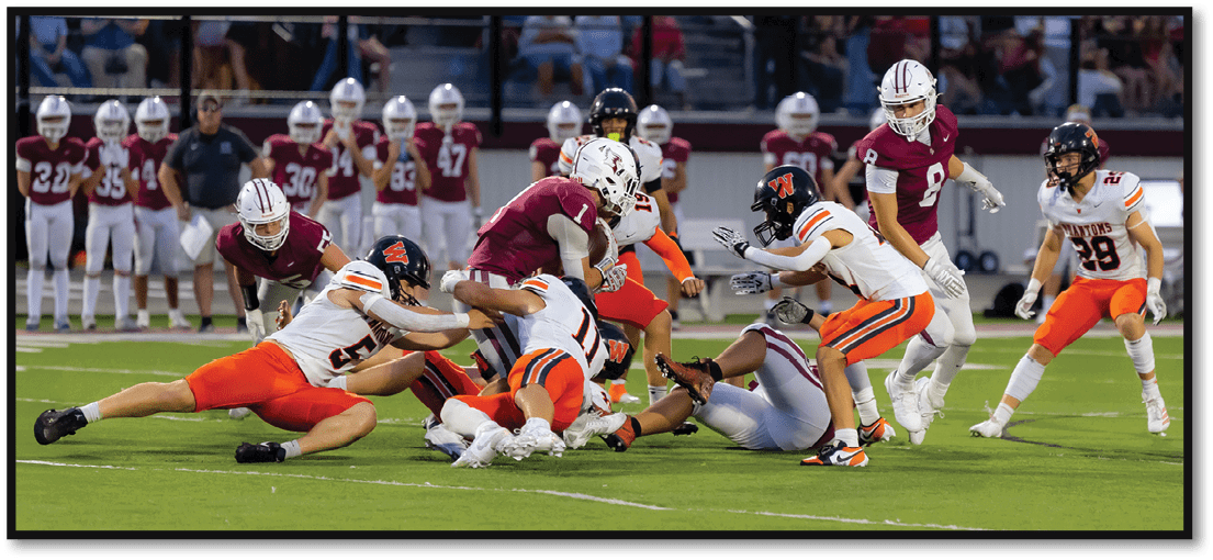 A group of people wearing football uniforms are actively engaged in a play on a football field with a crowd watching from the stands in the background. Description generated by AI